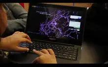 This is a picture of an open laptop on a desk.  The student using the laptop is not pictured but you can see one hand on the keyboard and one hand on the mouse pad as if they are in the middle of using the computer.  The website on the laptop says EyeWire in rainbow colors at the upper left of the screen and there is a menu option bar across the top of the webpage.  The webpage has a black background with a large picture of what appears to be a neuron structure (which looks like branches of purple squiggly lines coming from a small, spherical component).  To the right of the screen is where you enter login information and the top left it says, "What is EyeWire? Play a game to map the brain."