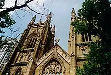 View looking up between Plane trees towards two towers with a gable between them. The towers and gable have panelling to match the window tracery and curvilinear ornamentation.