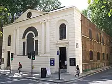 A large white building, dominated by a single large arched window. Another side of the building, faced in brick, recedes into the distance along a narrow street.