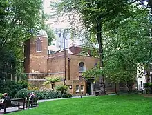 A patch of clear grass stands in front of a large red brick church with a distinctive narrow white bell tower. The church is surrounded by tall trees.