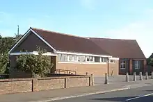 A long, low brick building in two non-aligned parts. Nearer the camera, in three-quarter view, is a hall with white soffits and window-frames; the windows are immediately below the roofline. Behind it, in profile and mostly hidden, is a longer building of brown and red brick and with a steep, high roof.