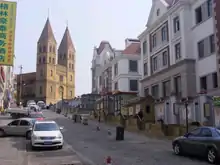 A street with Saint Michael's Cathedral rising in the distance. Cars are parked along one side of the street, and a row of four story buildings stand along the other side.