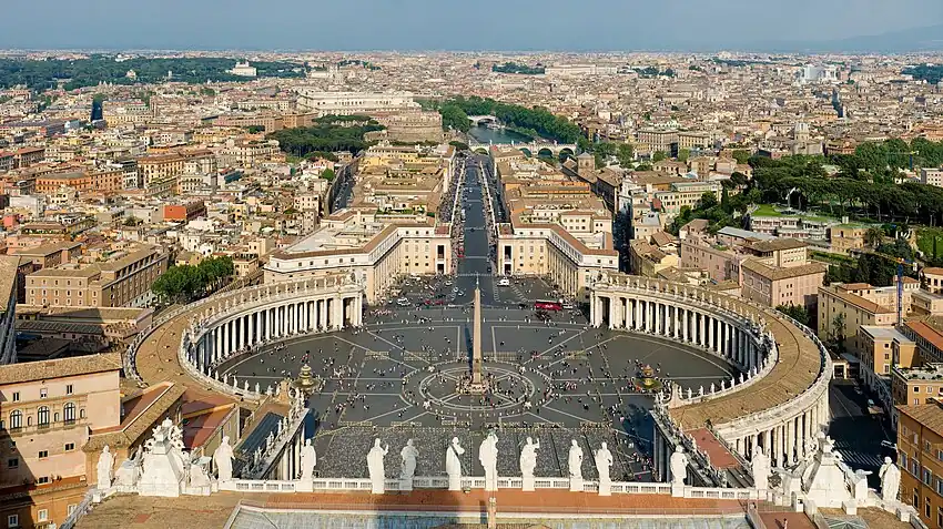 At the front of the view are the backs of thirteen large statues that stand in along the edge of the façade. Beyond them can be seen the piazza which is in three parts. The nearest appears square, while the second widens into an oval surrounded on each side by the huge grey columns on the colonnade, and with the obelisk at its centre. Beyond that is a further square surrounded by pale pink buildings. A wide street leads from the square, at the end of which can be seen the river, a bridge and castle.