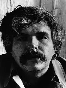 Close-up black-and-white portrait photograph of a man with wavy medium-length hair and a Walrus moustache