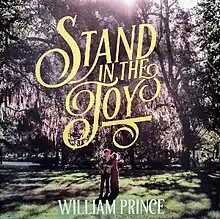 Picture of William Prince and his fiancée stood in a forest with the yellow text "Stand in the Joy" above them in ornate script.