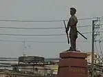 Side on view of a cement statue depicting a man with a moustache holding a rifle in one hand and a stick in another, wearing traditional clothes. He stands on a polished stone pedestal, and a city landscape and buildings can be seen in the background.