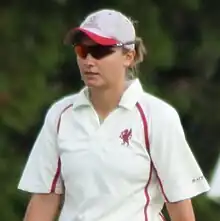 A woman wearing a white cricket t-shirt with red piping and a red dragon logo on the chest.  She is wearing sunglasses and a grey and red cap, under which her light brown hair is up.