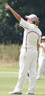 A woman with light brown hair wearing white cricket uniform with red piping points with her left arm.  Another player is visible in the background.