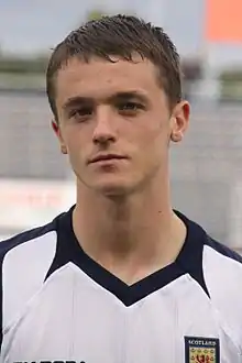 A man wearing a white shirt with a navy blue collar. The word "SCOTLAND" is printed on a badge on his chest