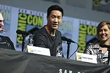 An Asian man with short black hair smiles towards the camera while sat behind a desk. A woman with short blonde hair sits next to him laughing. There are microphones on the desk in front of them.