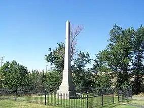 White stone obelisk surrounded by grass and a black fence