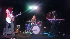 A shot of four women on stage in mid-performance. The first woman at right is cradling her guitar without playing it, she stands looking to her left: towards the second woman. She wears over shoulder-length red-brown hair, a white t-shirt with grey and black paisley-styled long pants and black shoes. The second woman is seated at her drum kit with her arms at her lap behind a drum. A microphone in a stand is above her. She wears slightly shorter hair, which is blonde-light brown, her shirt has a white background with a brown and grey design, and jeans. The third woman is partly obscured, she is playing her bass guitar and is turned to her left. She wears longer dark brown hair, a dark red-brown shirt with three-quarter sleeves over dark pants. The fourth woman is behind her keyboard and a microphone on a stand. The keyboard has writing: "nord electro 4". She stands turned to her right, facing the second woman. He arms are crossed with hands clasped at her waist. He legs are almost crossed, too. Her hair is shorter than the previous woman but has a similar colour. Her t-shirt is dark blue with jeans and dark shoes. Surrounding the women is stage equipment including speakers, instruments, electrical cords and a pair of stage lights (in the centre above the second woman).