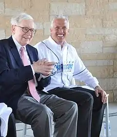 A man in a dark suit sitting beside a man wearing a white baseball jersey