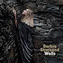 Barbra Streisand wears a black shall, gazing upward, surrounded by a large stone wall