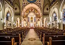 Interior view from the rear of the center aisle of a large, well lit, neo-Gothic church, toward the colorful apse.