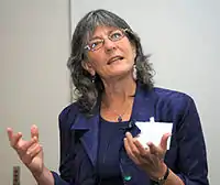 a woman in glasses stands looking up and to her left while holding her hands in front of her