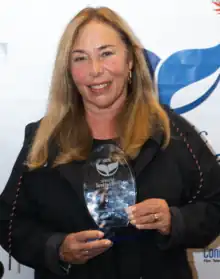 Picture of a middle-aged blonde woman. She's wearing a black sweatshirt and holds a crystal trophy with engraved letters to the camera. She stands in front of a white background with blue symbols and letters.