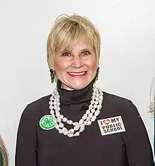 A white woman with blonde hair is wearing a brown turtleneck and a large white necklace; she is looking and smiling into the camera.