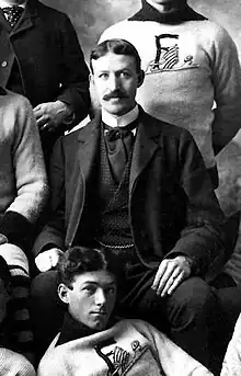 Black and white photo of hockey players seated and standing around Sutherland dressed in a suit and tie