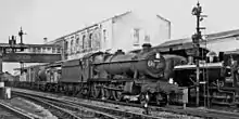 In a black and white photograph, a locomotive (seen from the front and right) is pulling a freight train, of which can be seen a number of open mineral wagons, a closed wagon, and some tankers. The driver is standing at the edge of the cab and seems to be looking at the photographer. The number 4936 is shown on the smoke box at the front, and also the number 681 is written in chalk. Behind the train there is a two-storey brick building which includes a covered bridge over the railway. To the right of the locomotive is a second, smaller, tank engine.