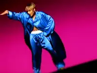 A women showing a white boxer inside her pants, with her right hand up and her left hand on her hip, backed by a dark pink background.