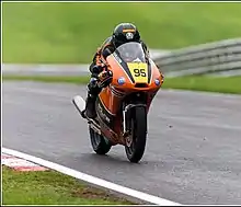 Racing motorcycle with brown bodywork and rider tucked in on smooth tarmac with a green backdrop of parkland