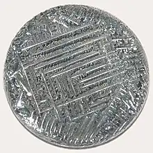 A shiny silver-white medallion with a striated surface, irregular around the outside, with a square spiral-like pattern in the middle.