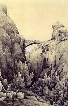 The entrance to a gorge in a rocky landscape, with a ruined bridge at the top and vegetation below