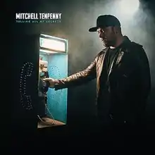 A man wearing a black t-shirt, leather jacket and baseball hat next to a payphone.