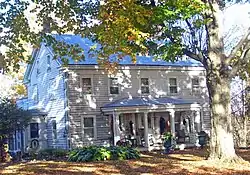 A white house with small front porch and a large tree in front. There are dead leaves on the lawn and some of the tree's leaves have turned yellow.