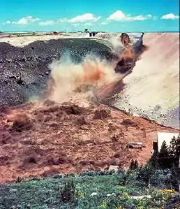 The dam is now breached and muddy water flows violently over the dam face.