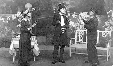 stage scene with a man in full mourning costume centre, woman to his right and man in clerical garb to his left