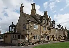 The Jubilee Tree stands on a small patch of grass, the tree is surrounded by a bench and behind it is an old farm building terraced with other houses.