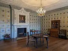  A sitting room decorated with panels painted with a 3D effect, containing a few pieces of furniture and a portrait by Thomas Gainsborough hanging over the fireplace.