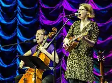 Photograph of two women singing and playing instruments on stage.