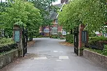 A gated entrance lined with trees leads to a red bricked building with white windows and a dark roof.