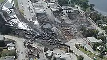 An aerial view of a small town on a lake. Over half the buildings are burnt to the ground, next to over 60 oil tanker cars derailed off a long sweeping curve. White foam covers the ground while firefighters cool the wreck.