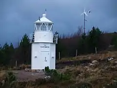 A white lighthouse on a hillside, with a wind turbine in the background