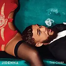 Jidenna rests his head on a woman's knee as he blows a puff of smoke.