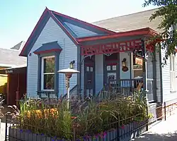 A small light blue wooden house with dark blue, and in some cases red, trim. There is a small plot with grassy plants and flowers in front, and signs saying "Matsuhisa".