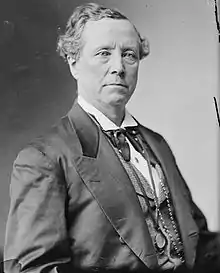 A man with wavy, receding hair, wearing a black suit with a white shirt, seated