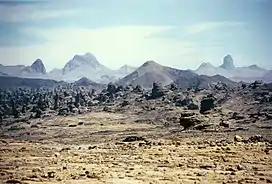 A hazy-looking yellow field of desert sand with rocks in the foreground and dark mountains and the blue sky in the background