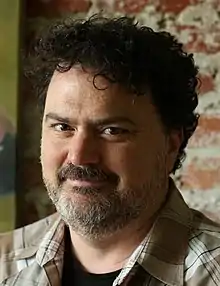 A Caucasian man in a brown plaid shirt looks into the camera, standing against a wall of brick.