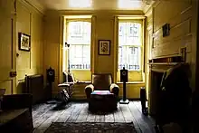 A wood panelled room, with polished wood floor, partly covered by a rug in the foreground. An armchair is between two sash windows, through which are the tops of spiked metal railings and, further away, Georgian brick houses with similar windows. On the mantlepiece above the fireplace are colourful spools of silks. In the foreground is a dummy, dressed in formal jacket, shirt and tie.