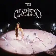 Tini sitting in the middle of a pink circle, surrounded by arrows lodged in the ground. The arrows have heart shaped tips. The backround is black and above Tini is her name and the album title, which is printed in a pink swirling font.