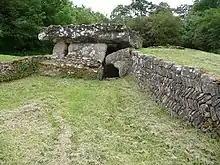 a grassy field in which two low stone retaining walls flank a stone structure with a thick stone slab roof