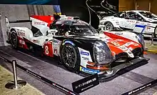 Side view of a red, white and black Toyota TS050 Hybrid enclosed in a space at a automobile show