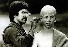 Savini, on the left, applies make-up behind Lehman's ear. Lehman's bald head has been made to appear over-large; his eyes point in  directions, and his teeth are extremely crooked.