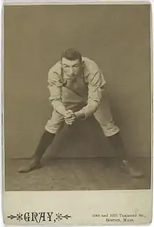 A black-and-white photograph of a man in a white baseball uniform bent over from the waist but looking into the camera