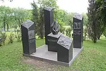 A black stone headstone of Richard in the foreground, atop a grass field, with trees in the background